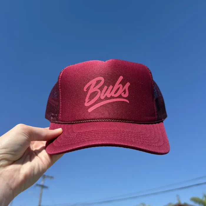 Hand holding a BUBS Burgundy Trucker Hat outdoors