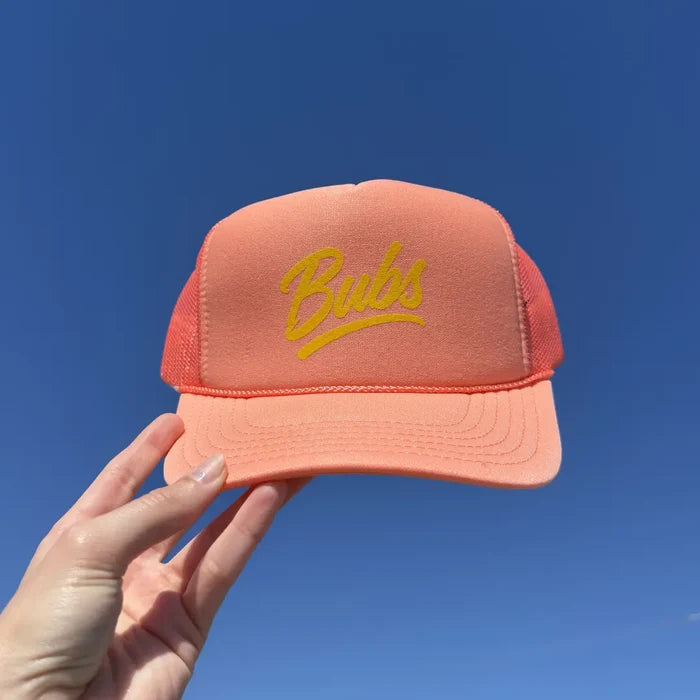Hand holding a BUBS Coral Trucker Hat outdoors