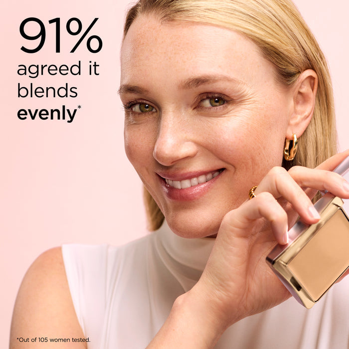 Woman applying makeup with a gold compact, smiling, on a pink background.