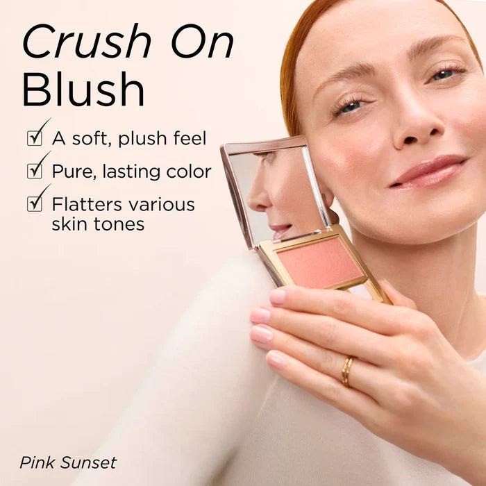 A woman holds a compact blush mirror showing a pink blush pan next to her cheek, with text reading 'Crush On Blush' and 'Pink Sunset' on a soft beige background.