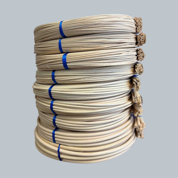 A stack of coiled natural rattan round reeds, each a different size, bundled with a white paper label, set against a solid gray background.