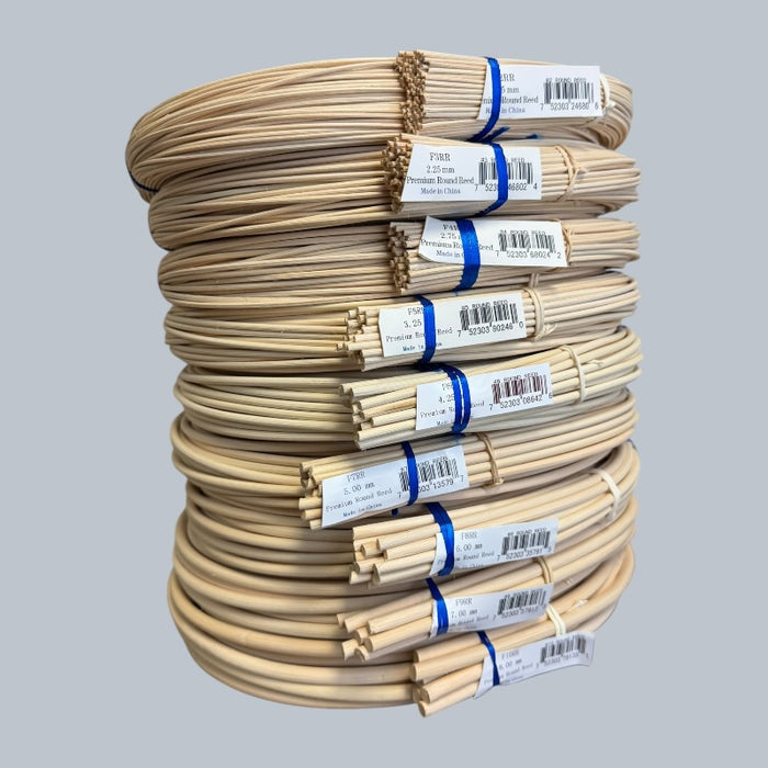 A stack of coiled natural rattan round reeds, each a different size, bundled with a white paper label, set against a solid gray background.